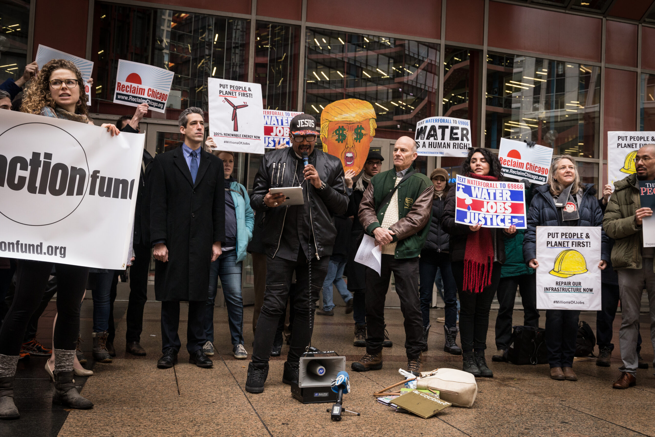 A person speaking into a mic surrounded by other people holding different signs in front of a building.