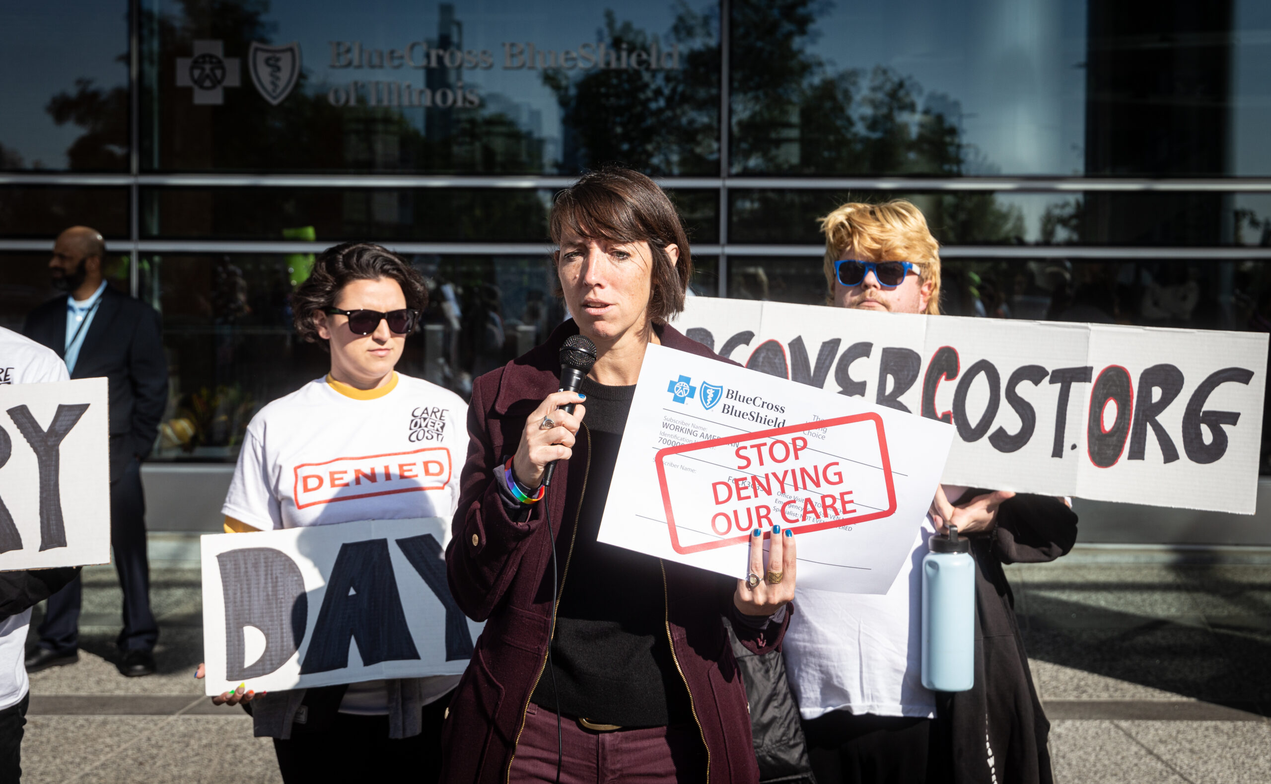 Person with a mic and a sign that says “Stop denying our care” and a few other protestors in front of a building.