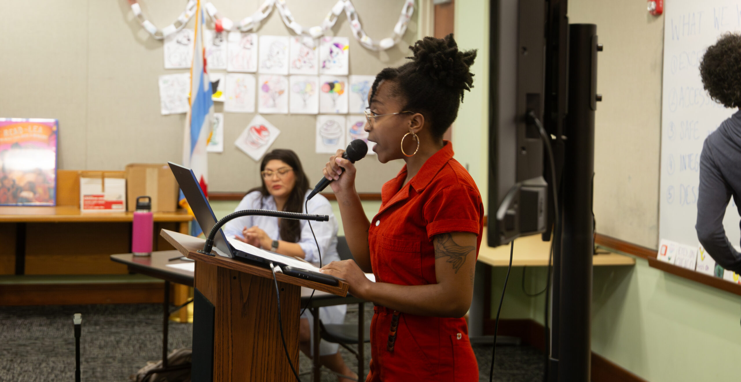 A person at a podium facing left and speaking into a mic in a classroom.