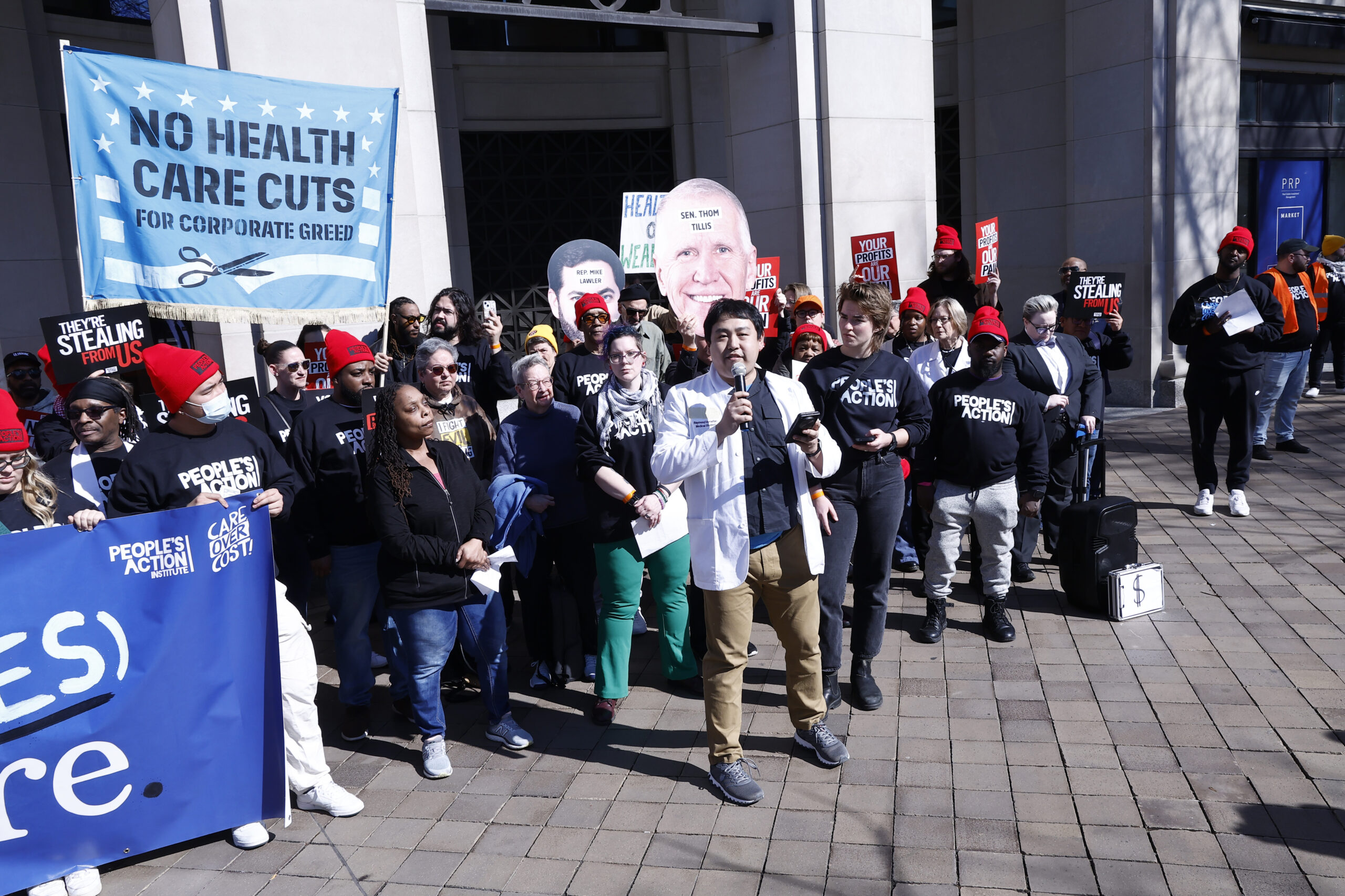 A group of protesters stand together, many wearing People's Action T-Shirts. Some hold a large sign saying, "NO HEALTH CARE CUTS."